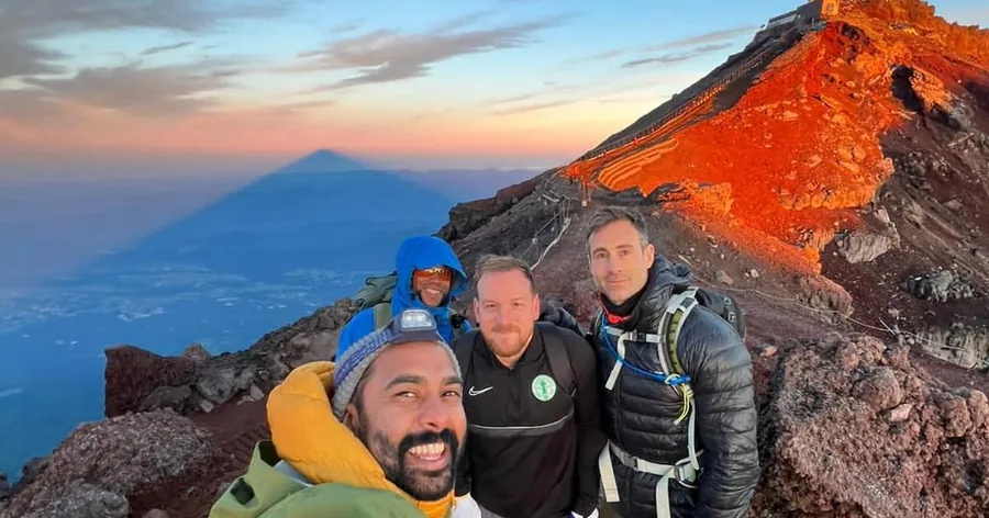 御来光の中、富士山の赤い火山斜面で笑顔を見せる3人の登山者。雲に富士山の影が落ちています